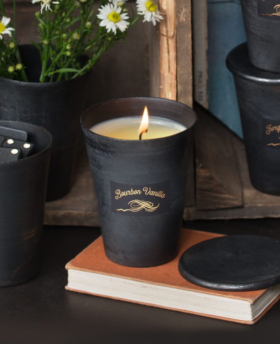 Candle in black cup holder with 'Bourbon Vanilla' label on a wooden surface with plants and books in the background