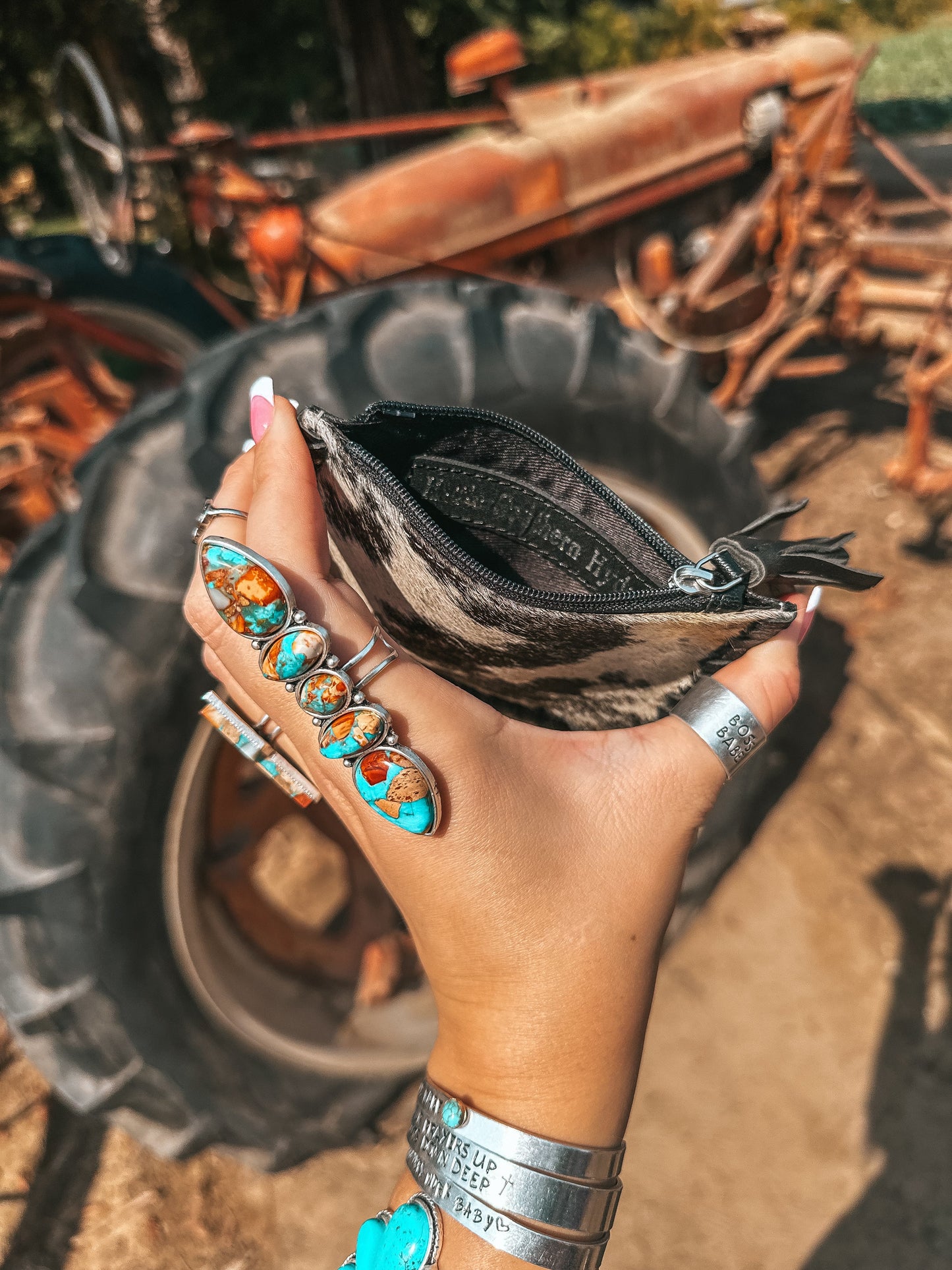 Hand holding a small black and white cowhide pouch with a rustic background featuring old tractors.
