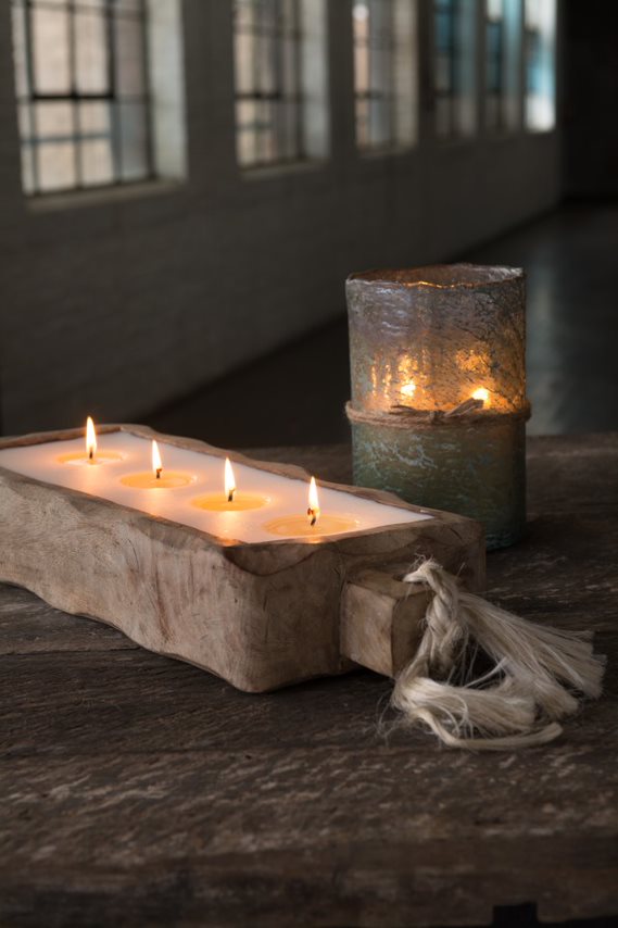 Wood carved candle holder with multiple wicks on a wooden table, next to a textured glass candle