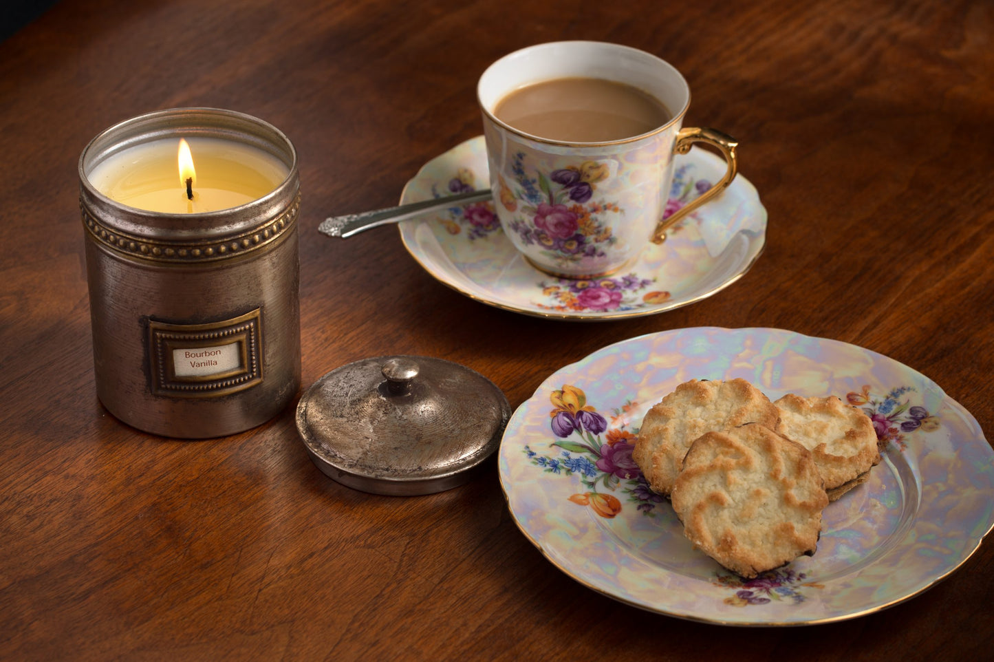 Candle in a decorative metal container, cup of tea on saucer with cookies on a plate all set on a wooden table