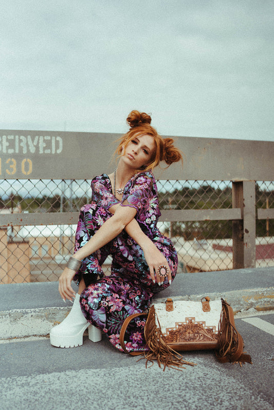 Woman in a floral dress sitting on a bench with a western handbag, outdoors.