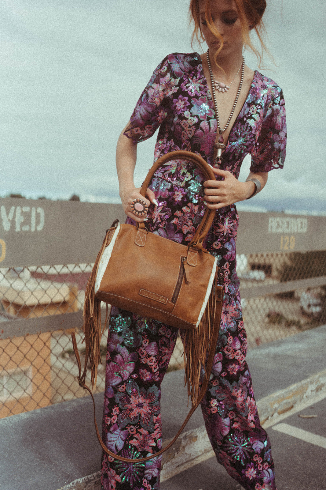 Person wearing a floral outfit holding a brown western leather handbag with fringe.