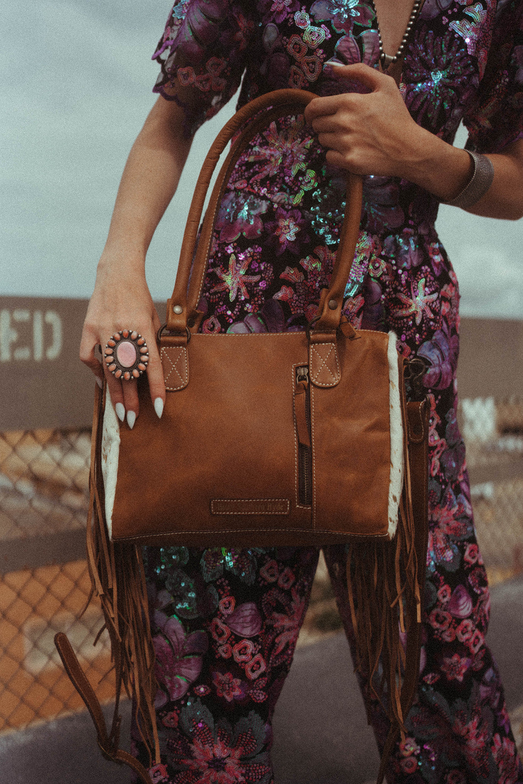 Person holding a brown leather handbag with fringe details, wearing a floral dress.