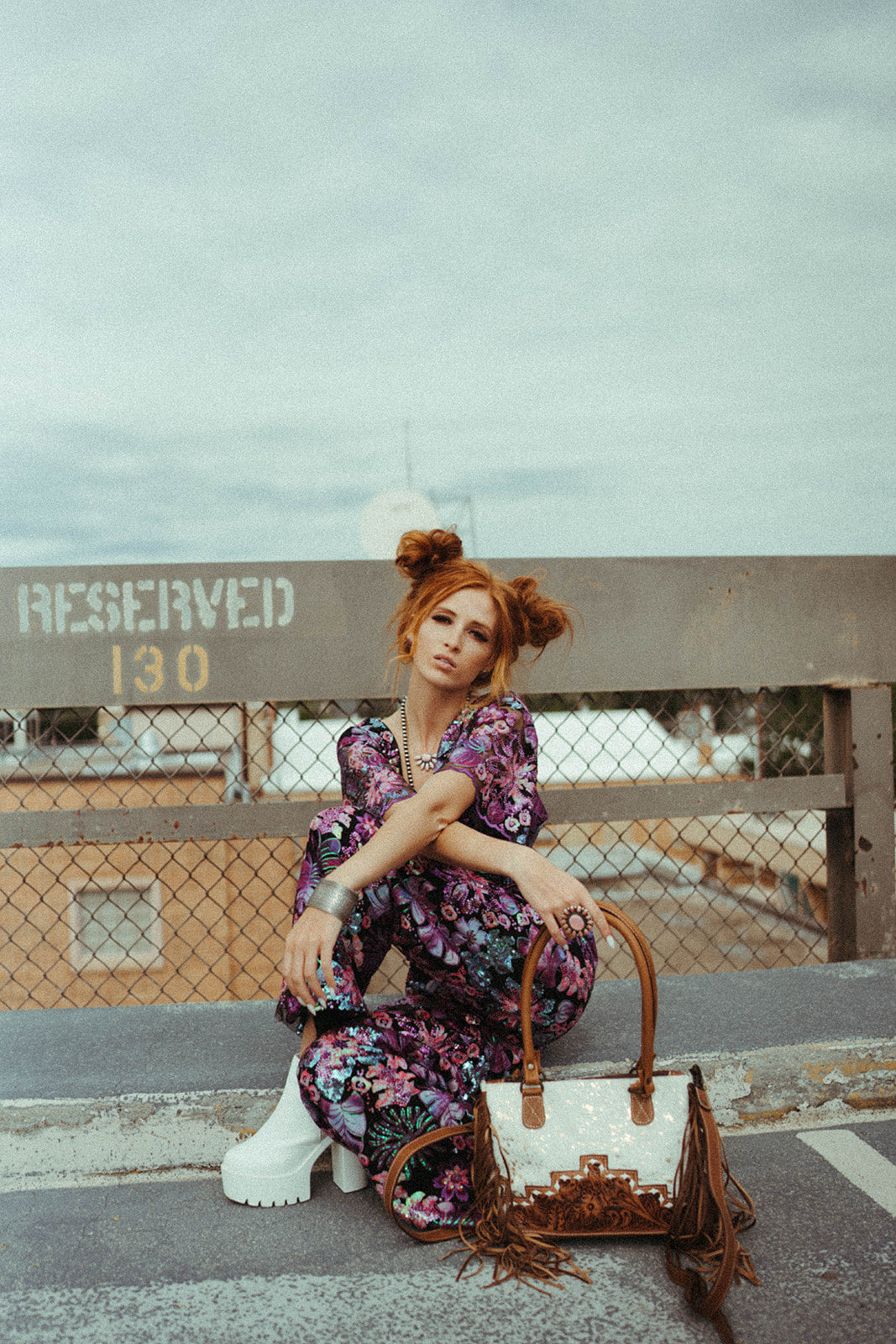 Woman in a floral dress sitting on a bench with a western fringed handbag, against a fence and sky background.