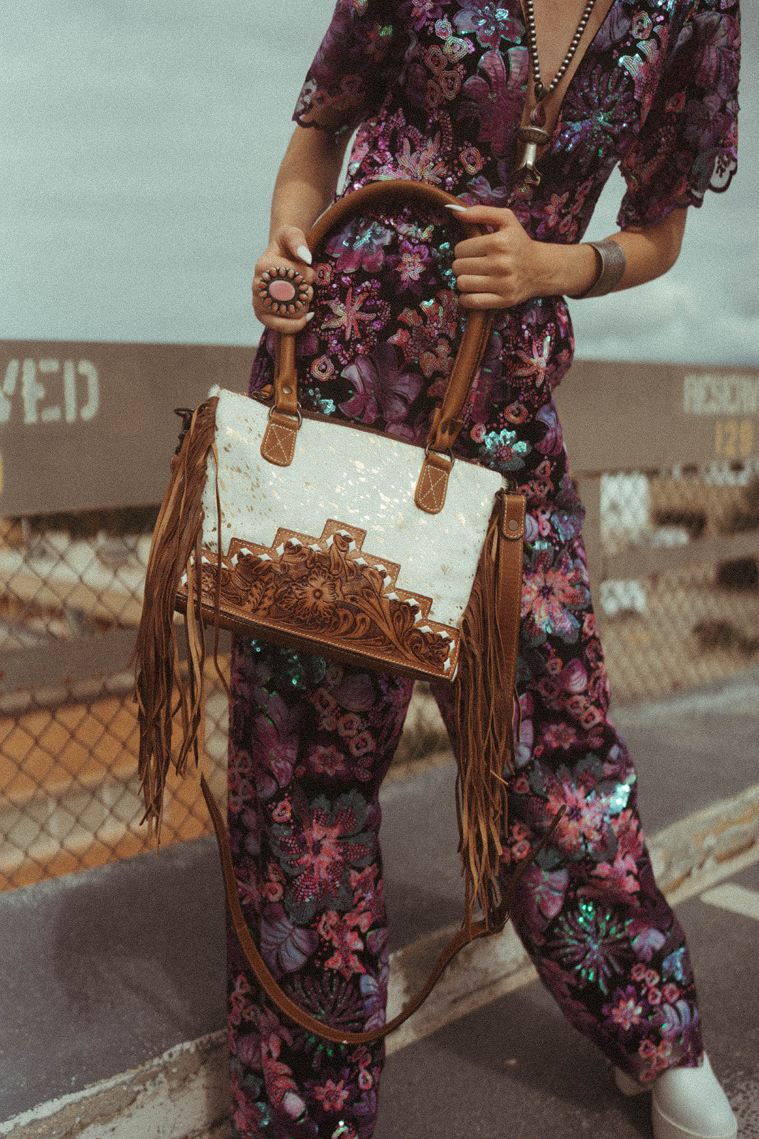 Woman wearing a floral outfit holding a decorative western handbag in an industrial setting.