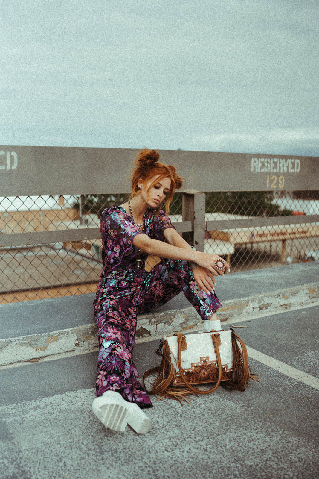 Woman in a floral jumpsuit sitting on a concrete surface with a western handbag beside her.
