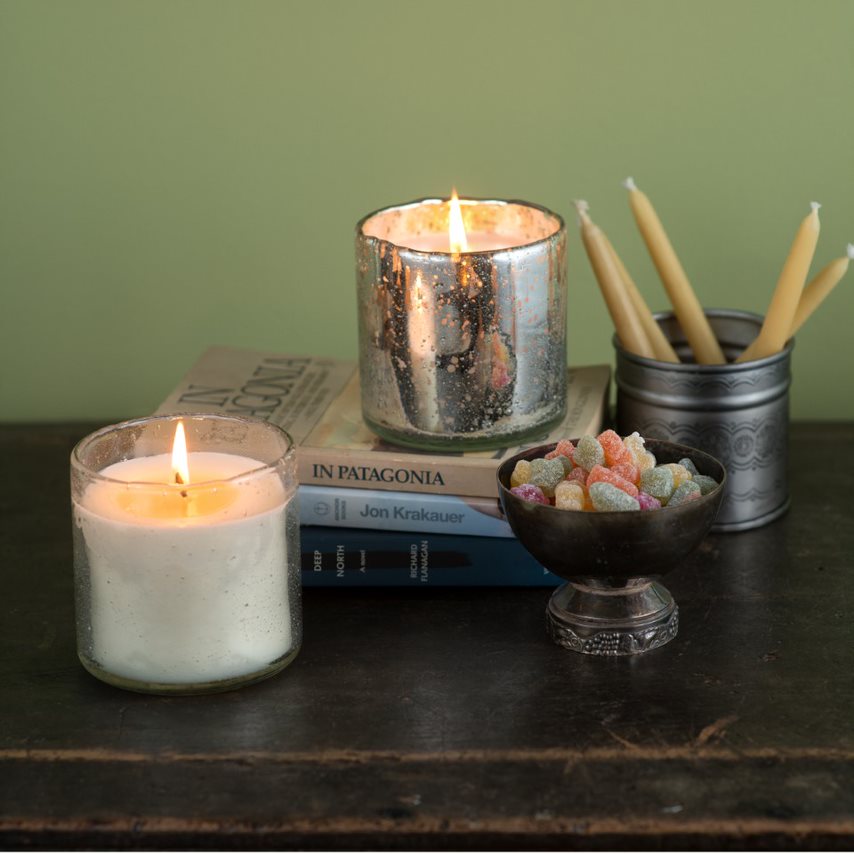 Two lit candles, a small bowl of colorful candies, and a book on a wooden surface with a green wall background.