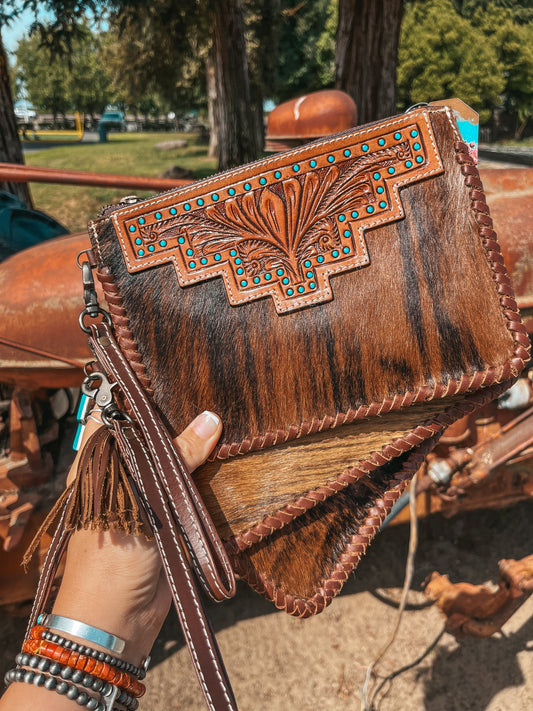 Brown leather clutch with intricate designs held by a person, with a blurred outdoor background.