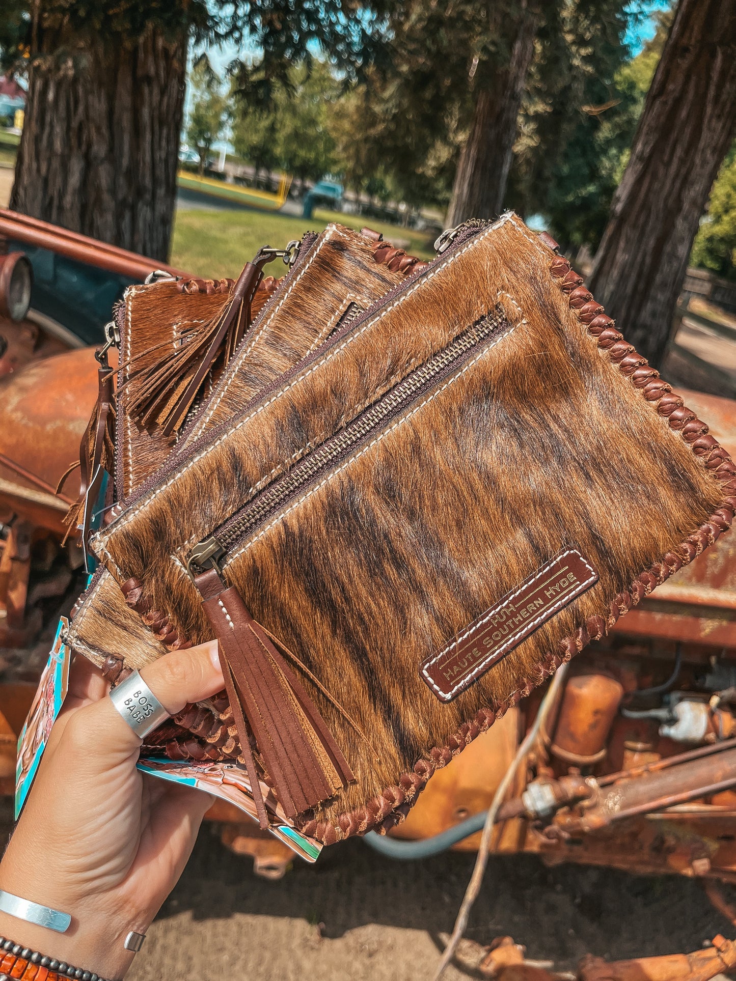 Brown leather clutch with fringe and brand logo held by a hand outdoors.
